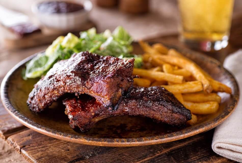Two pieces of barbecued ribs on a brown plate with fries and salad