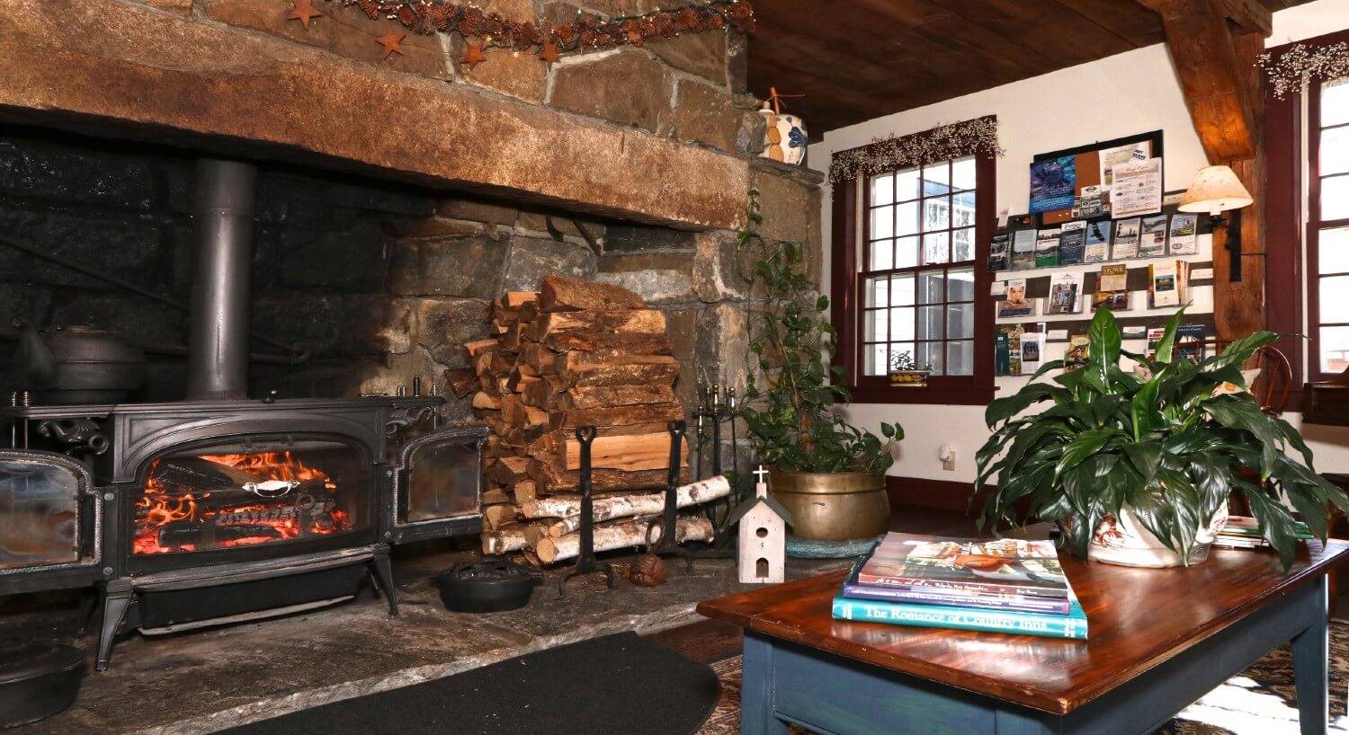 Large stone fireplace with wood burning stove, stack of firewood and coffee table with books and a plant