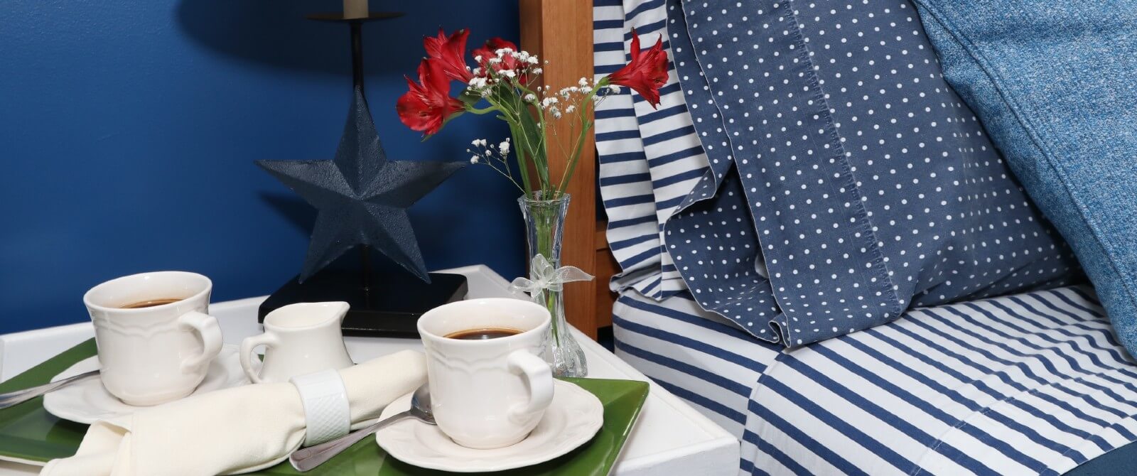 Bed with striped sheets and blue pillows with side table holding tray of coffee mugs and flowers