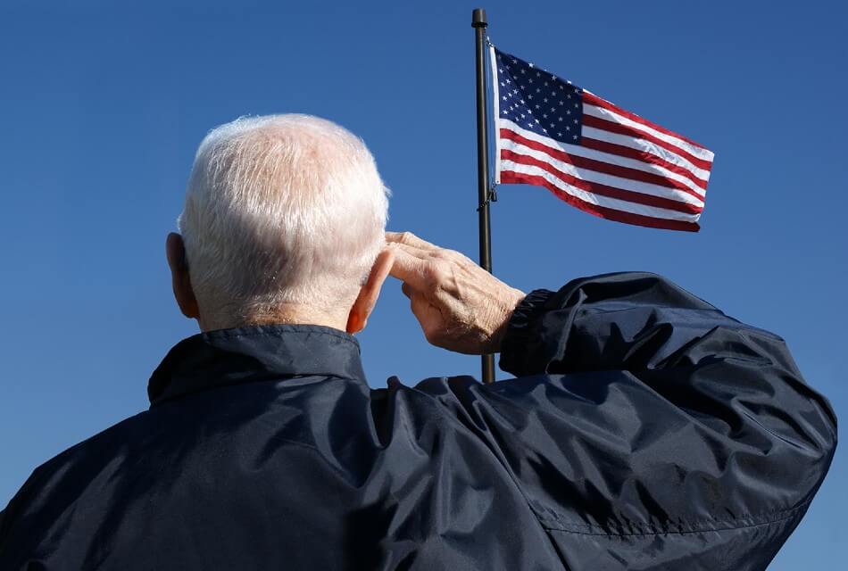 Older gentleman facing and saluting an American flag waving against a blue sky